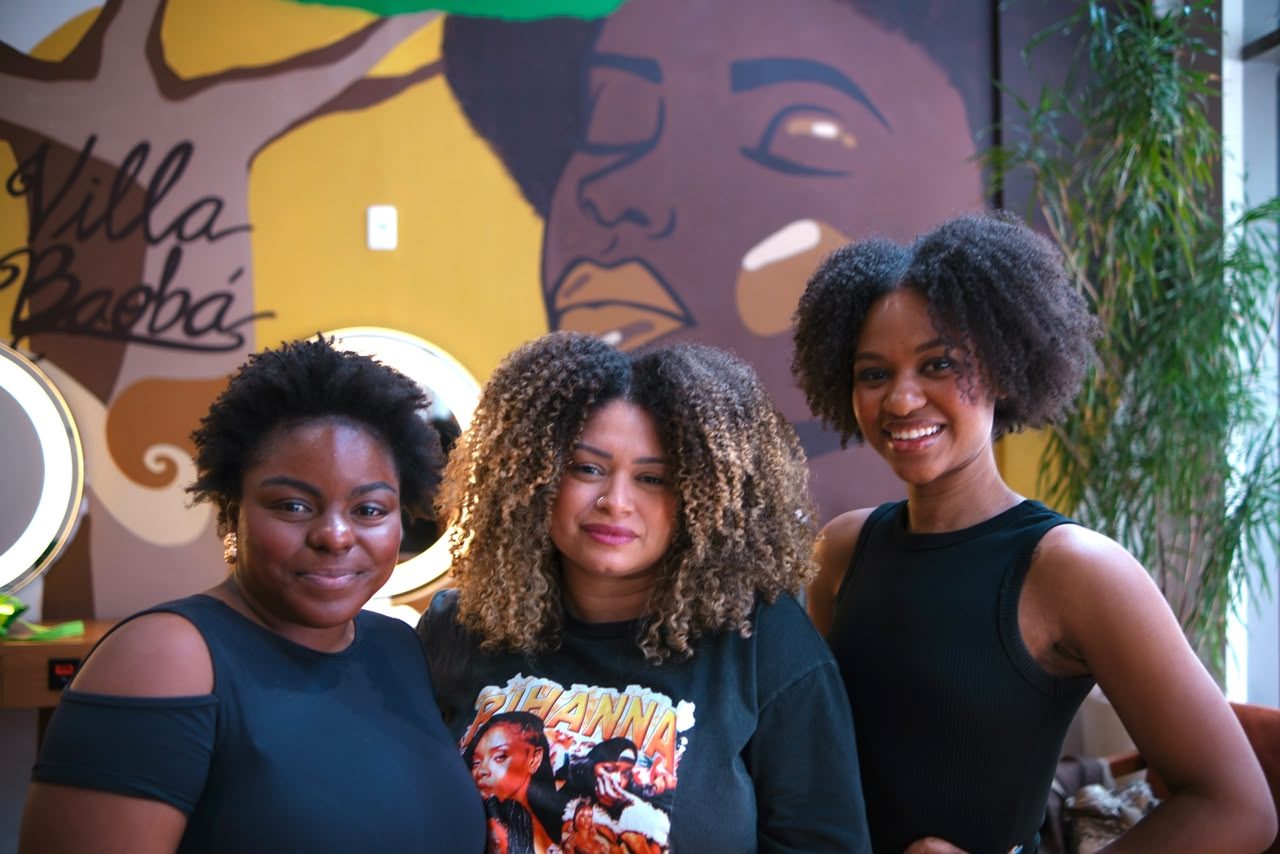 Três clientes sorrindo em frente ao mural Villa Baobá
